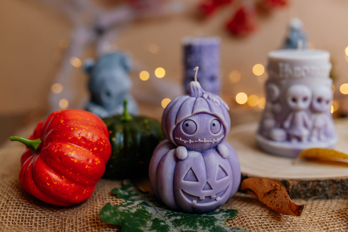 A purple carved jack-o'-lantern candle with a cute cartoon face sits on a burlap surface, styled with small pumpkins, autumn leaves, and other decorative candles in a warm, softly blurred setting.