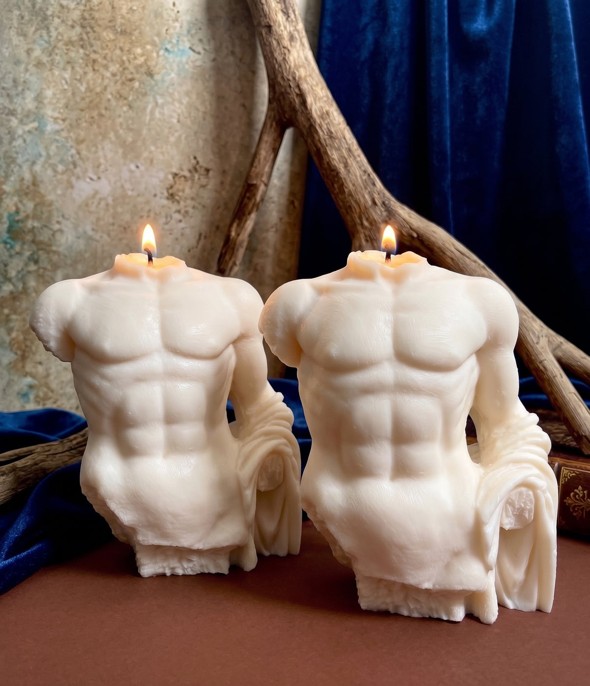 Two white wax candles shaped like male torso sculptures with visible muscular definition, shown lit on a brown surface with blue fabric and driftwood in the background.
