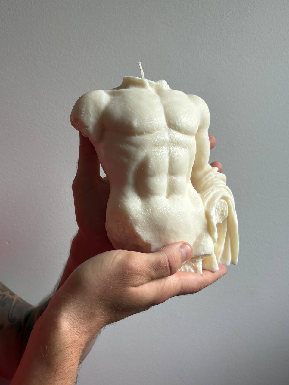 A hand holds a sculpted body-shaped candle designed to look like a muscular male torso, in an off-white or cream color, against a plain light gray wall.