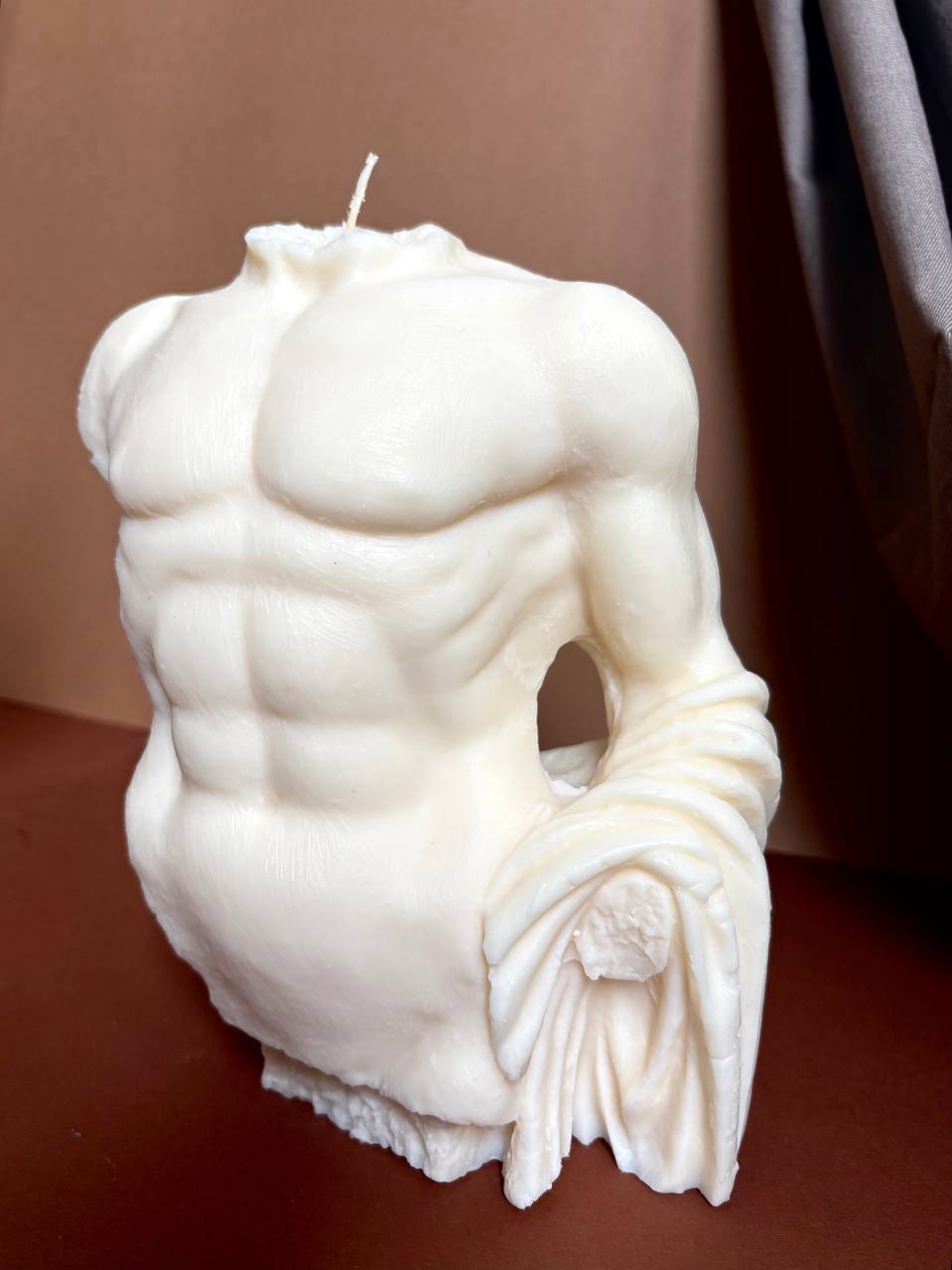 A white wax candle sculpted in the shape of a classical male muscular torso with draped fabric at the waist, shown on a brown surface against a brown backdrop.