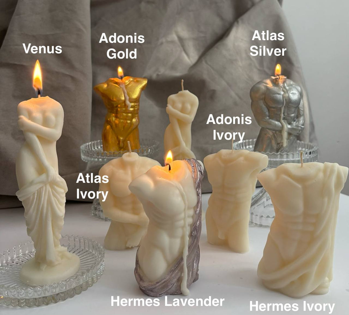 A group of sculptural body candles shaped like classical male and female statues, displayed on a white surface with a draped gray fabric backdrop. Visible labels include Venus, Adonis Gold, Atlas Silver, Adonis Ivory, Hermes Lavender, and Hermes Ivory.