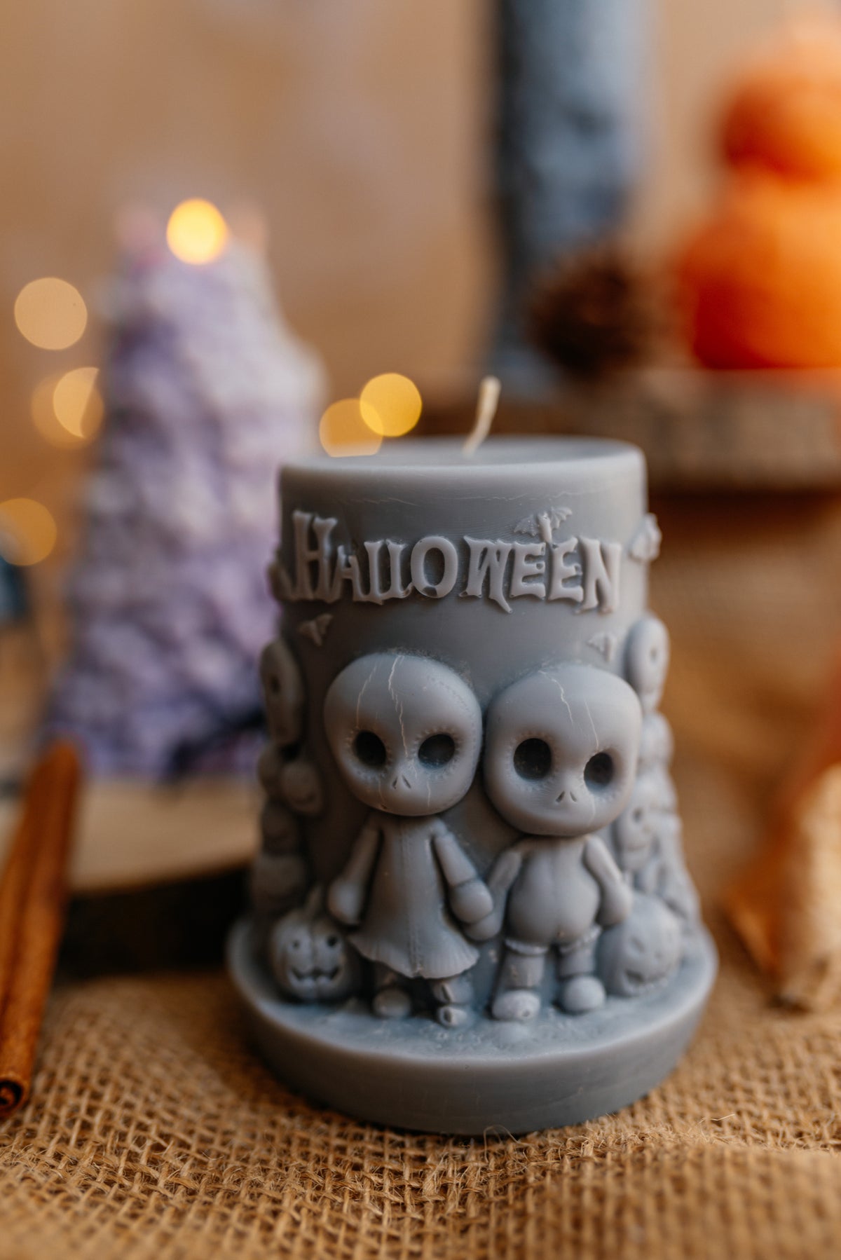 A gray Halloween-themed candle with two cute cartoon ghosts and the word 'HALLOWEEN' embossed on the front, displayed on a burlap surface with warm blurred background lights.