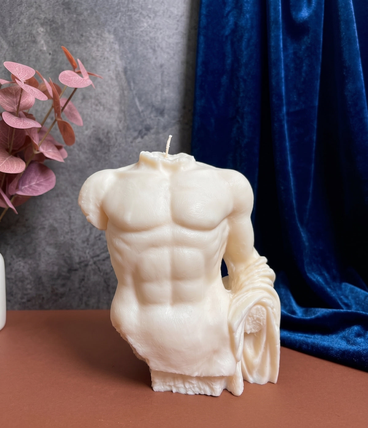 A sculptural candle shaped like a male torso with detailed muscular definition and draped fabric, shown on a tabletop with a gray textured wall, blue velvet drape, and pink leaves in the background.