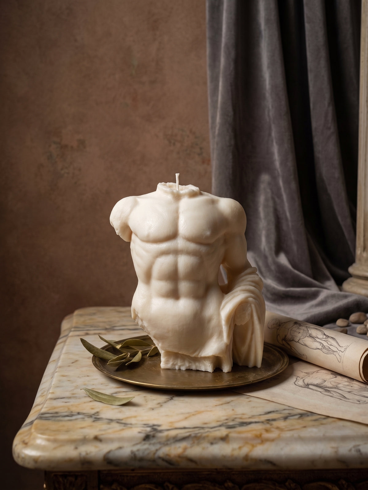 A sculptural male torso candle with detailed muscular definition, displayed on a round brass tray on a marble table with a rolled parchment and leaves against a textured brown wall and gray drape.