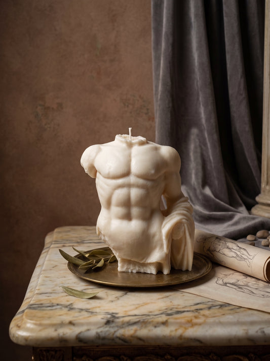A sculptural male torso candle with detailed muscular definition, displayed on a round brass tray on a marble table with a rolled parchment and leaves against a textured brown wall and gray drape.