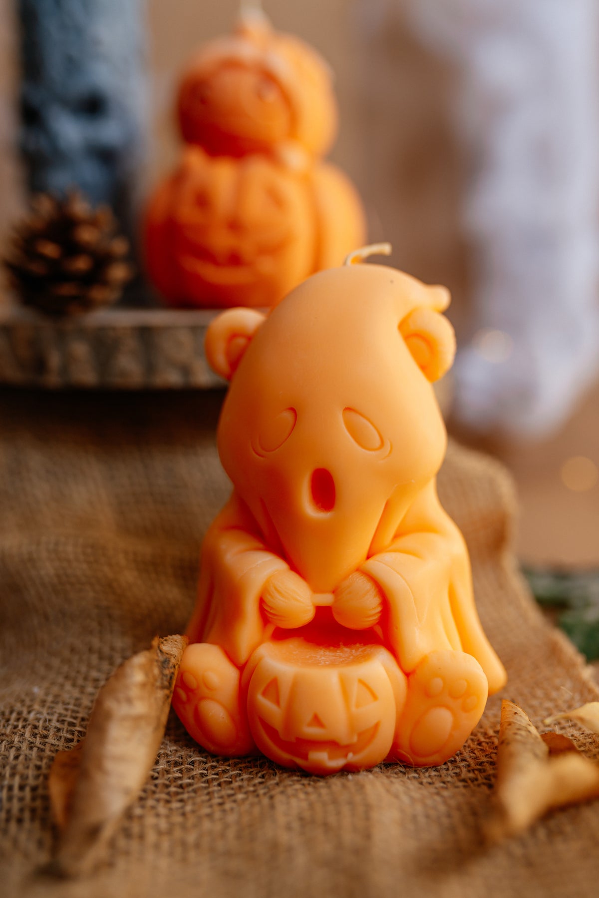 An orange Halloween-themed bear candle holding a carved jack-o'-lantern, styled on burlap fabric with a pinecone and a blurred matching candle in the background.