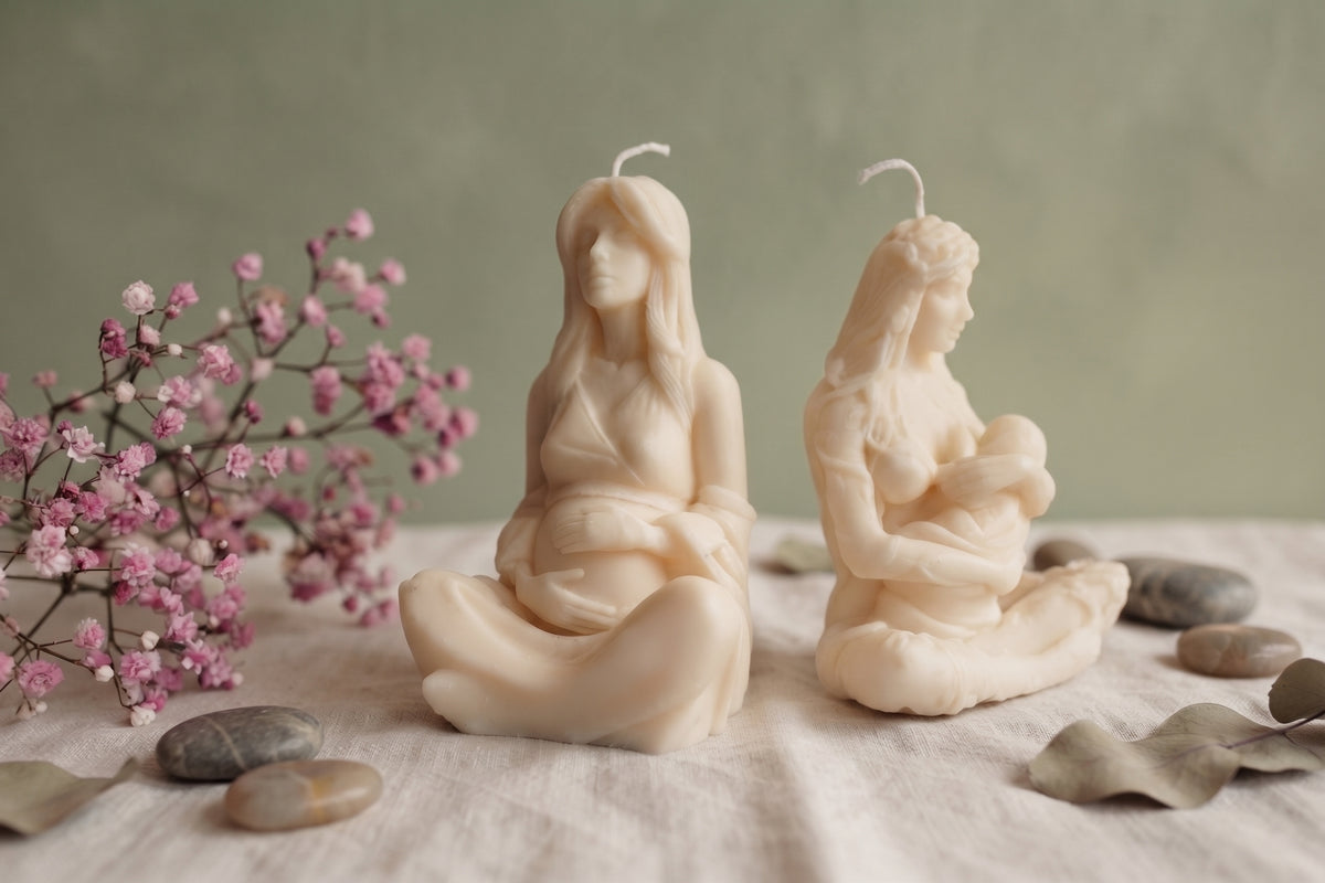 Two detailed cream soy wax candles in the form of female figures sit on a beige linen cloth. The left candle depicts a pregnant woman with hands on her belly. The right candle depicts a woman breastfeeding an infant. The arrangement is complemented by a sprig of pink baby's breath flowers (Gypsophila), smooth river stones, and dried eucalyptus leaves against a muted olive-green wall.
