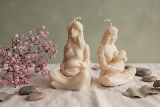 Two detailed cream soy wax candles in the form of female figures sit on a beige linen cloth. The left candle depicts a pregnant woman with hands on her belly. The right candle depicts a woman breastfeeding an infant. The arrangement is complemented by a sprig of pink baby's breath flowers (Gypsophila), smooth river stones, and dried eucalyptus leaves against a muted olive-green wall.