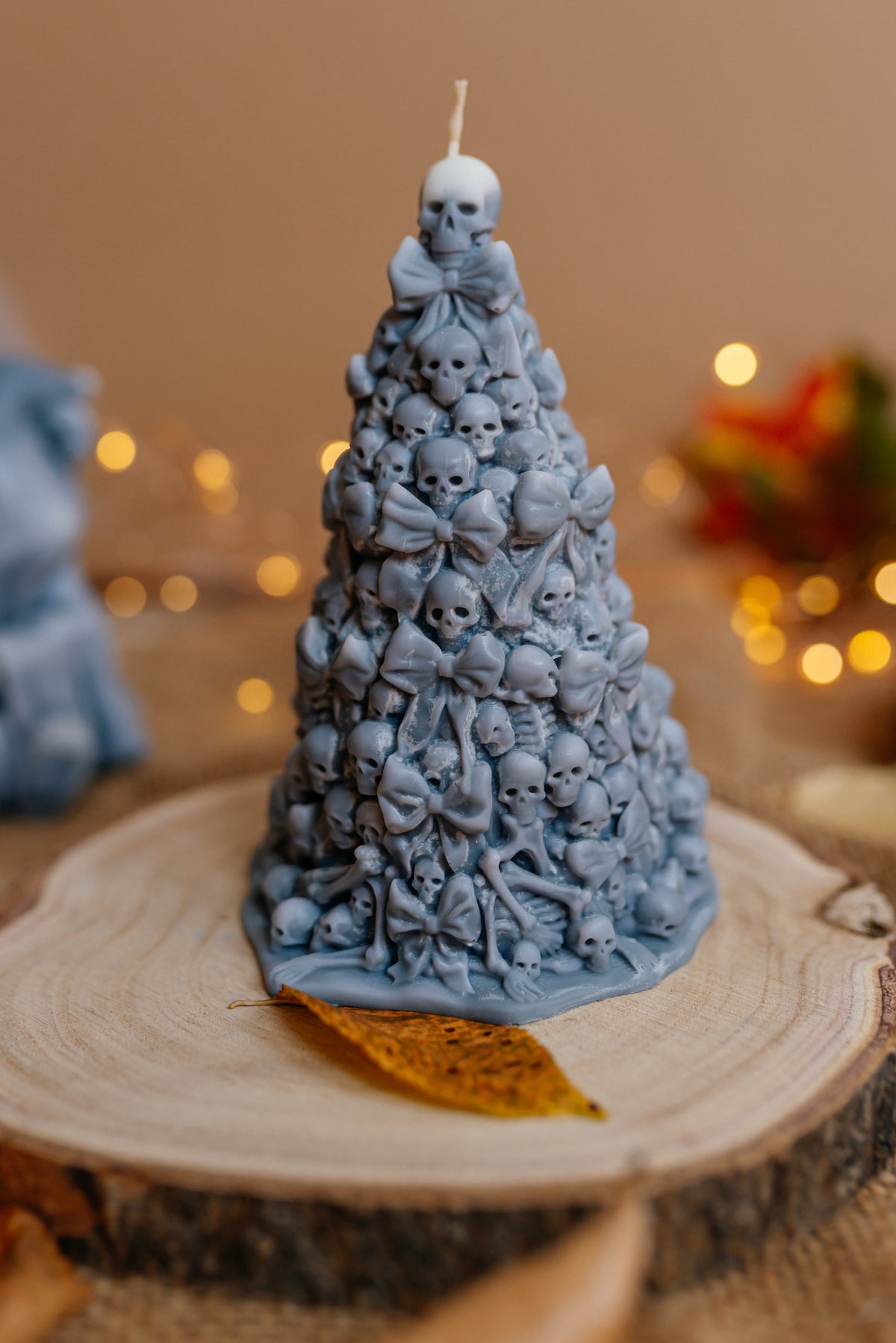 A grey Christmas tree-shaped candle covered in small skull and bow details, displayed on a round wood slice with warm blurred lights in the background.