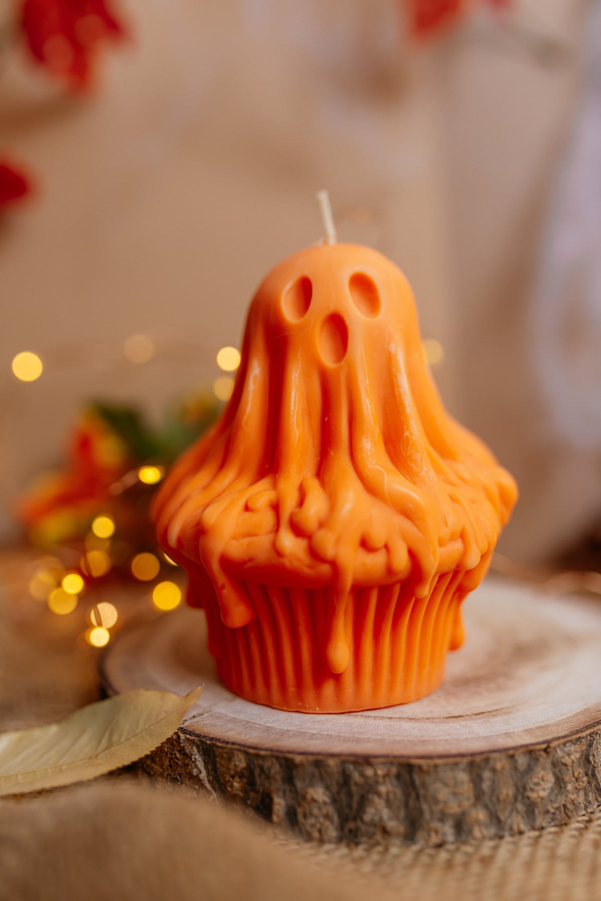 An orange Halloween ghost-shaped candle styled like a cupcake, sitting on a rustic wood slice with warm bokeh lights in the background.