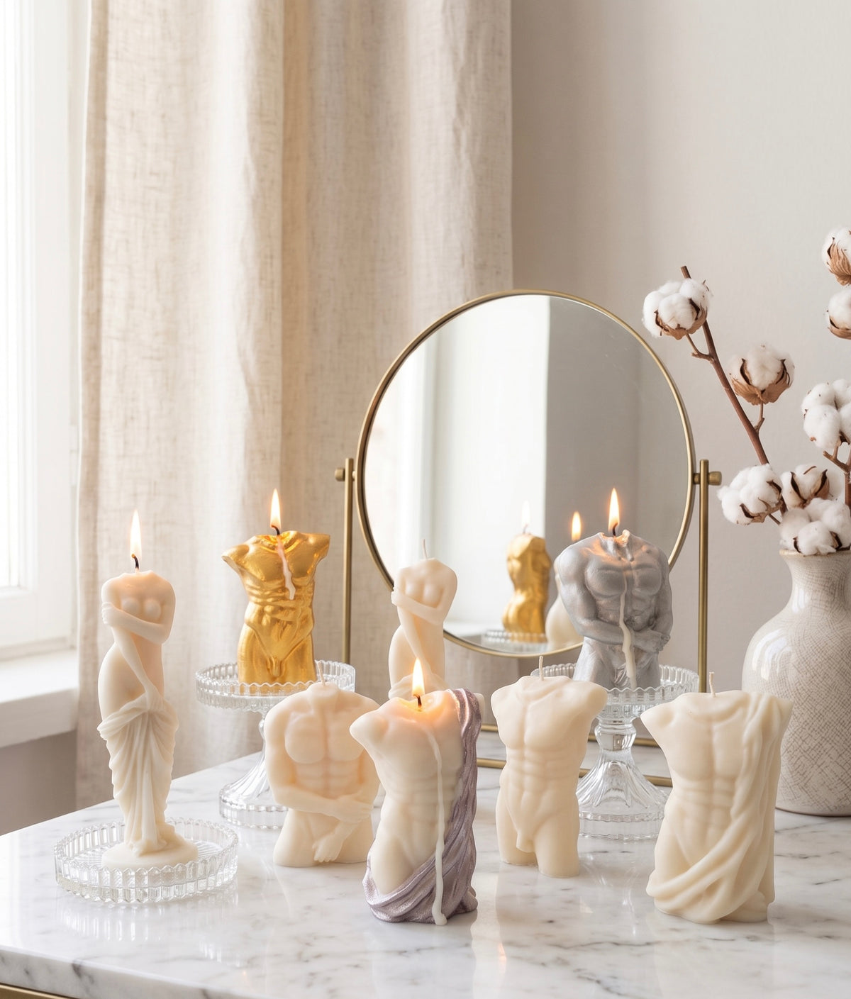 A collection of sculptural body-shaped candles in cream, gold, and gray, styled on a marble table with a gold-framed mirror, sheer curtains, and cotton bolls in a vase.