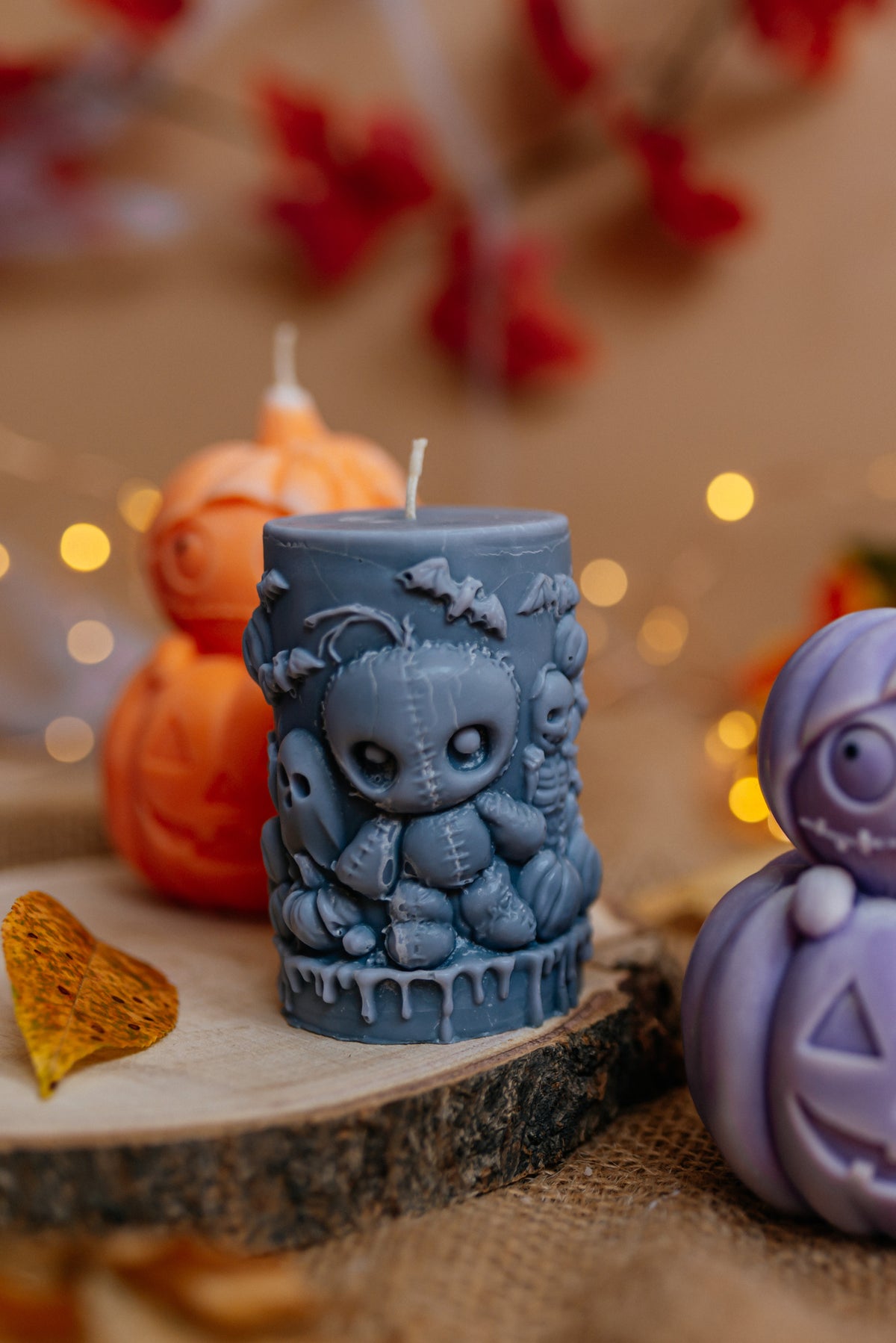 A dark gray Halloween candle featuring a stitched doll design, displayed on a rustic wood slice with blurred orange pumpkins and warm bokeh lights in the background.