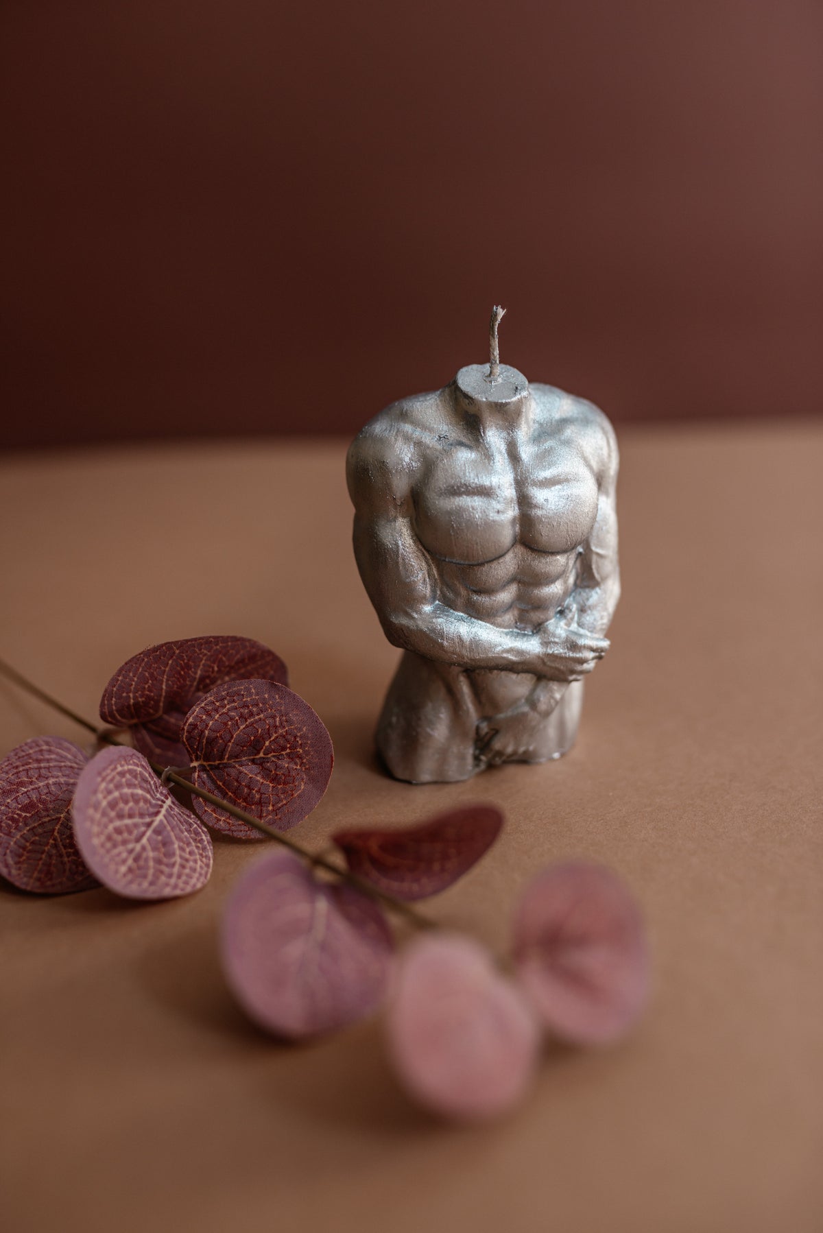 A sculptural muscular male torso candle in a metallic silver finish, styled on a brown surface with dried pinkish-brown leaves against a solid brown background.