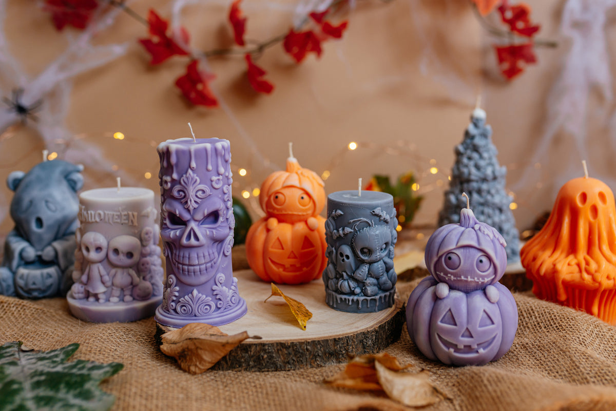 A group of Halloween-themed candles displayed on a burlap surface with autumn leaves and warm string lights in the background. The candles include purple skull, purple pumpkin, blue owl, orange jack-o'-lantern, blue ghost, and grey tree shapes.