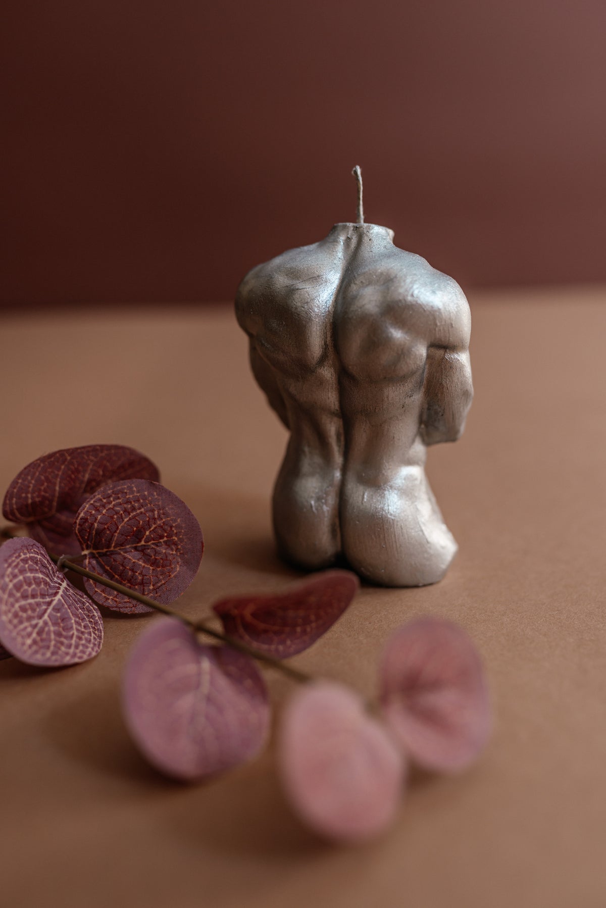 A sculptural candle shaped like a muscular male torso, shown from the back on a brown surface with blurred dried flowers in the foreground.