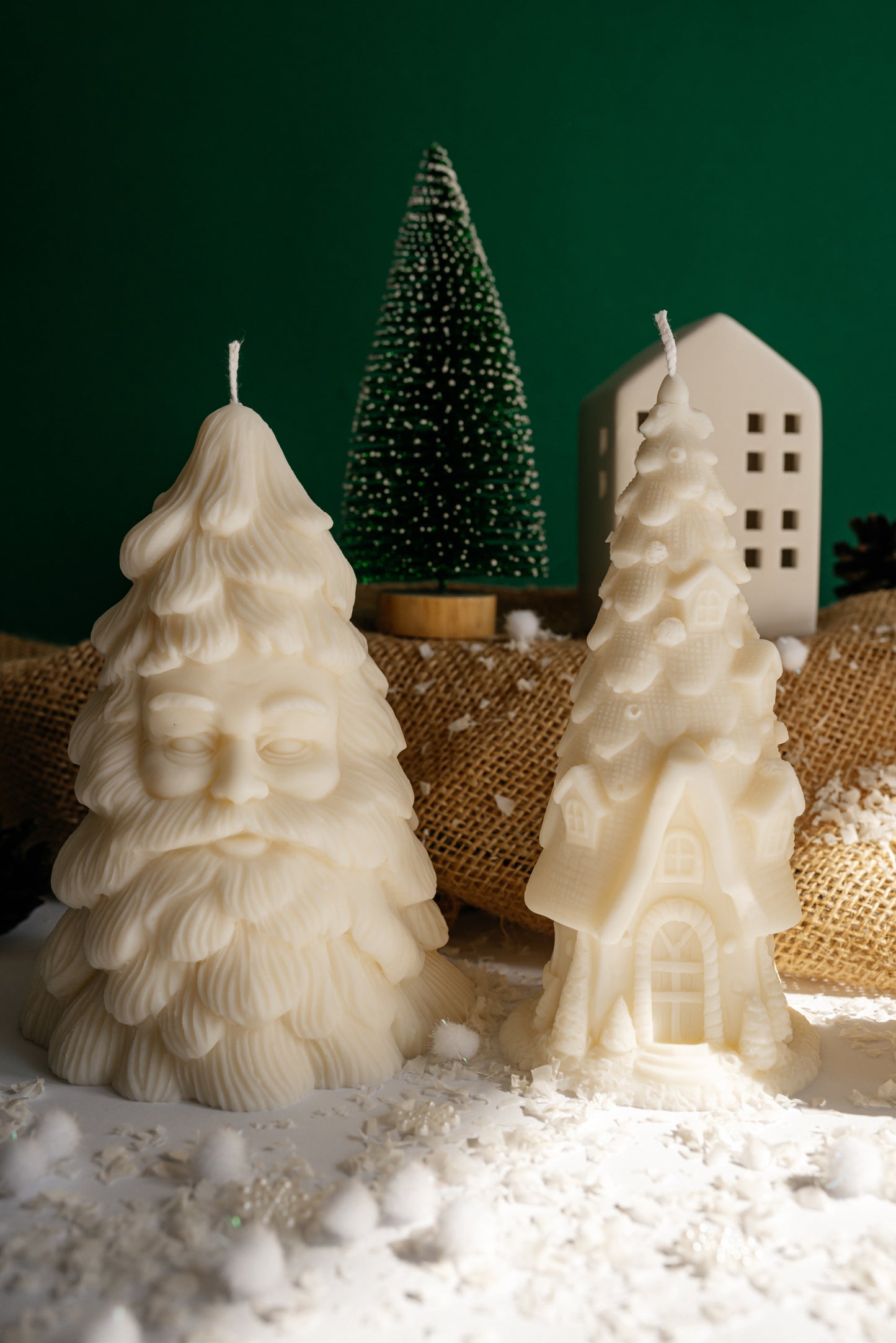 White soy wax candle shaped like a Santa Claus Christmas tree standing next to a house candle on festive snow background.