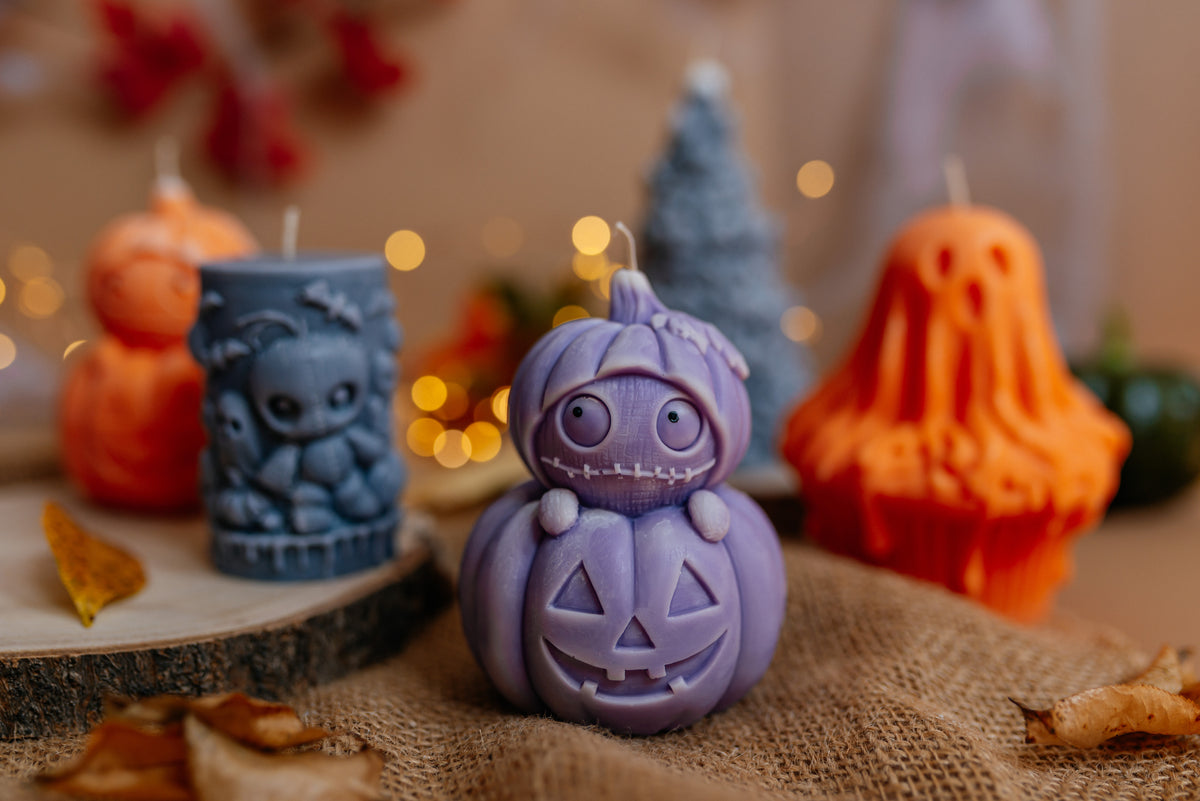A purple carved jack-o'-lantern candle sits on burlap in a styled Halloween setting, with orange ghost and pumpkin candles and a blue candle blurred in the background.