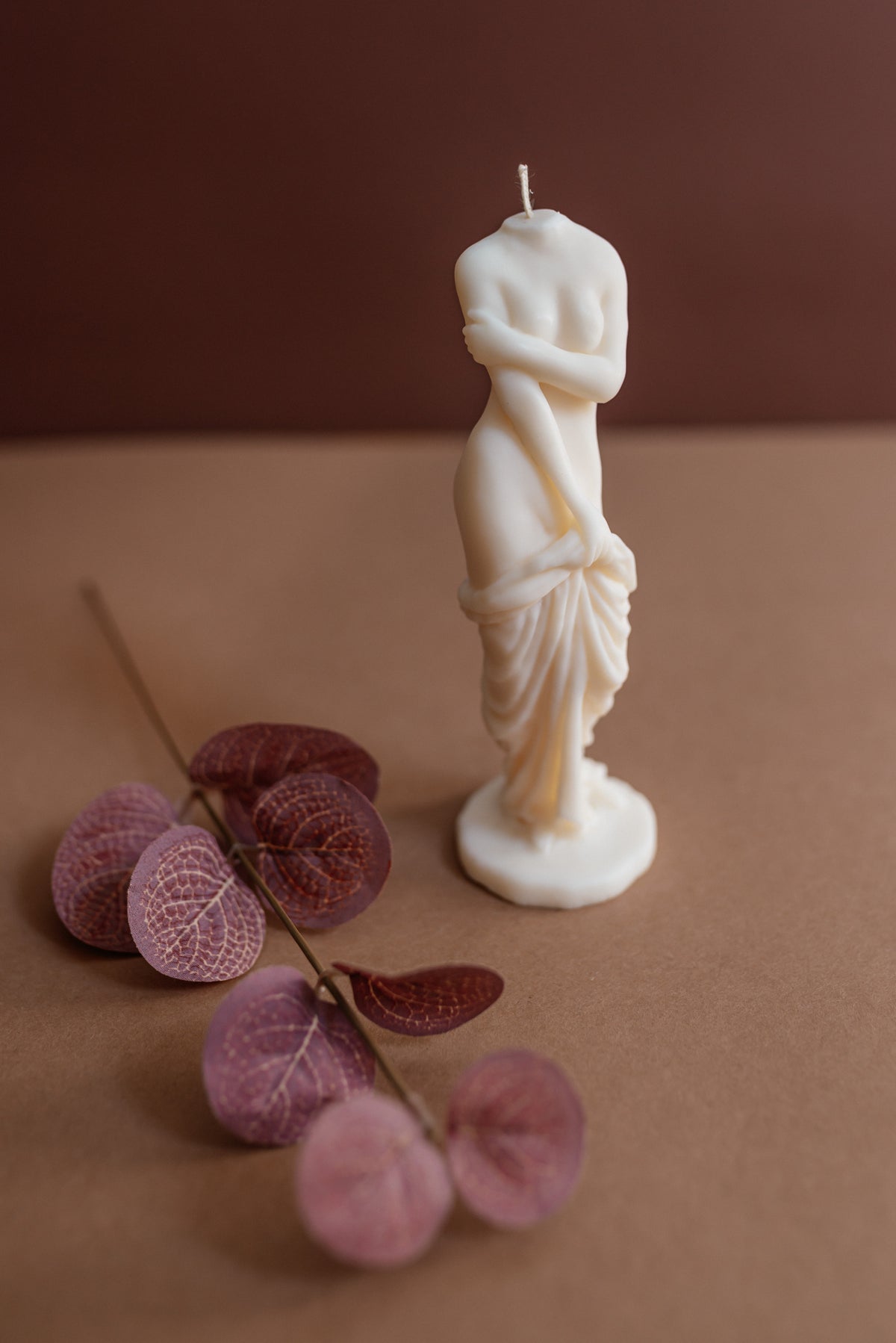 An off-white wax candle sculpted in the shape of a classical draped female statue, displayed on a brown surface next to dried pinkish-purple leaves against a solid brown background.