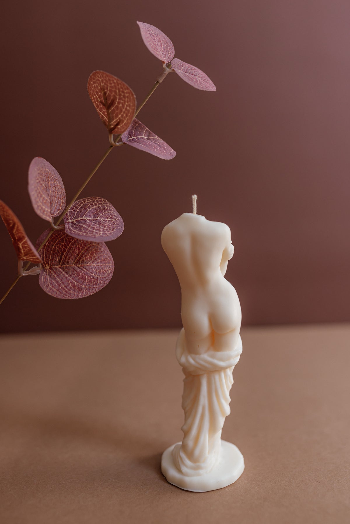 A cream-colored sculptural candle shaped like a classical female torso stands on a round base against a warm brown background, with dried pinkish-brown leaves on the left side.