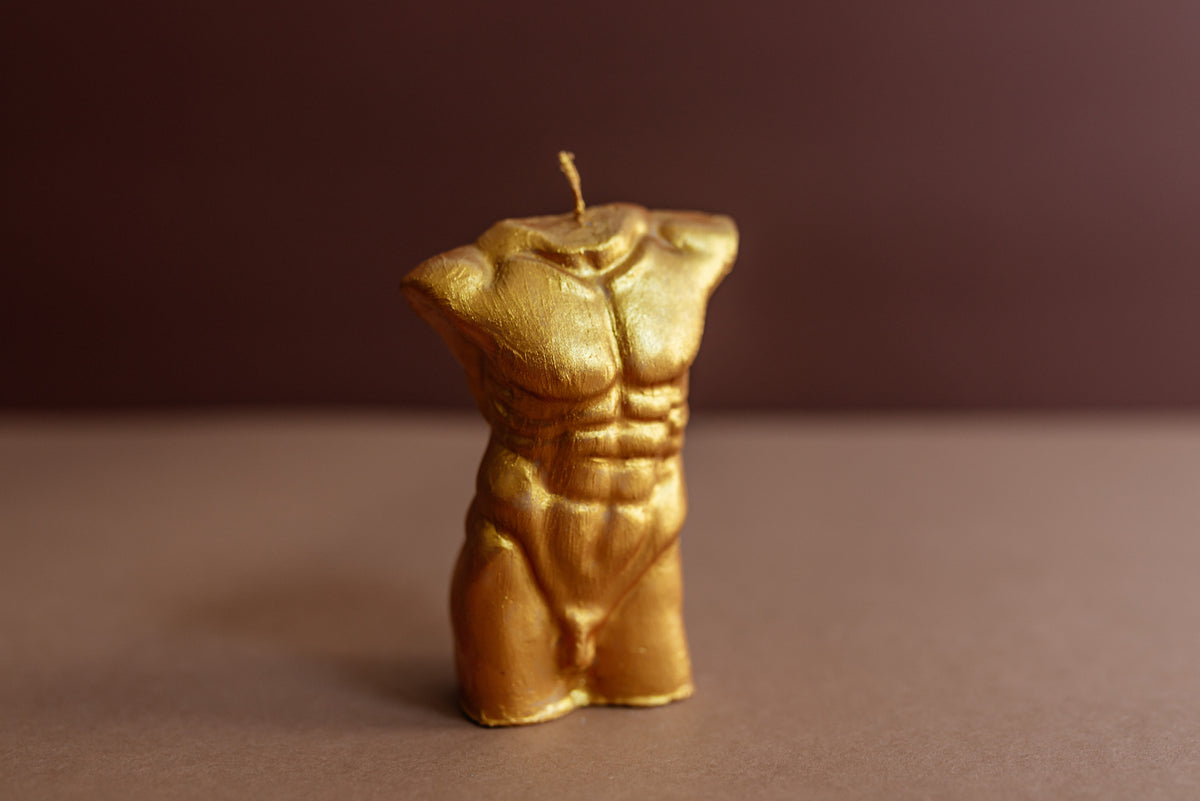 A gold-colored wax candle shaped like a male torso with detailed abdominal muscles, standing upright on a brown surface against a dark brown background.