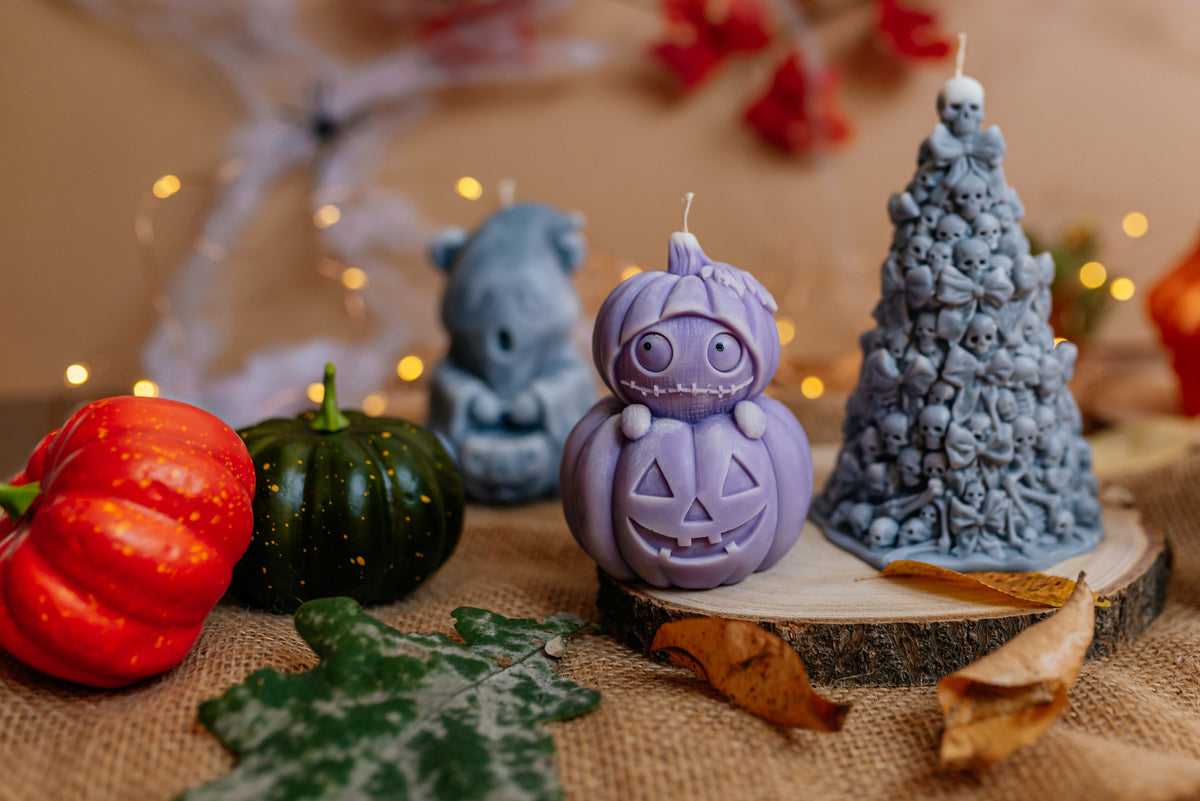 Halloween-themed candles displayed on a rustic wood slice, featuring a purple jack-o'-lantern candle, a blue Christmas tree candle with skull details, and a blue bear candle, surrounded by decorative pumpkins, autumn leaves, and warm string lights.