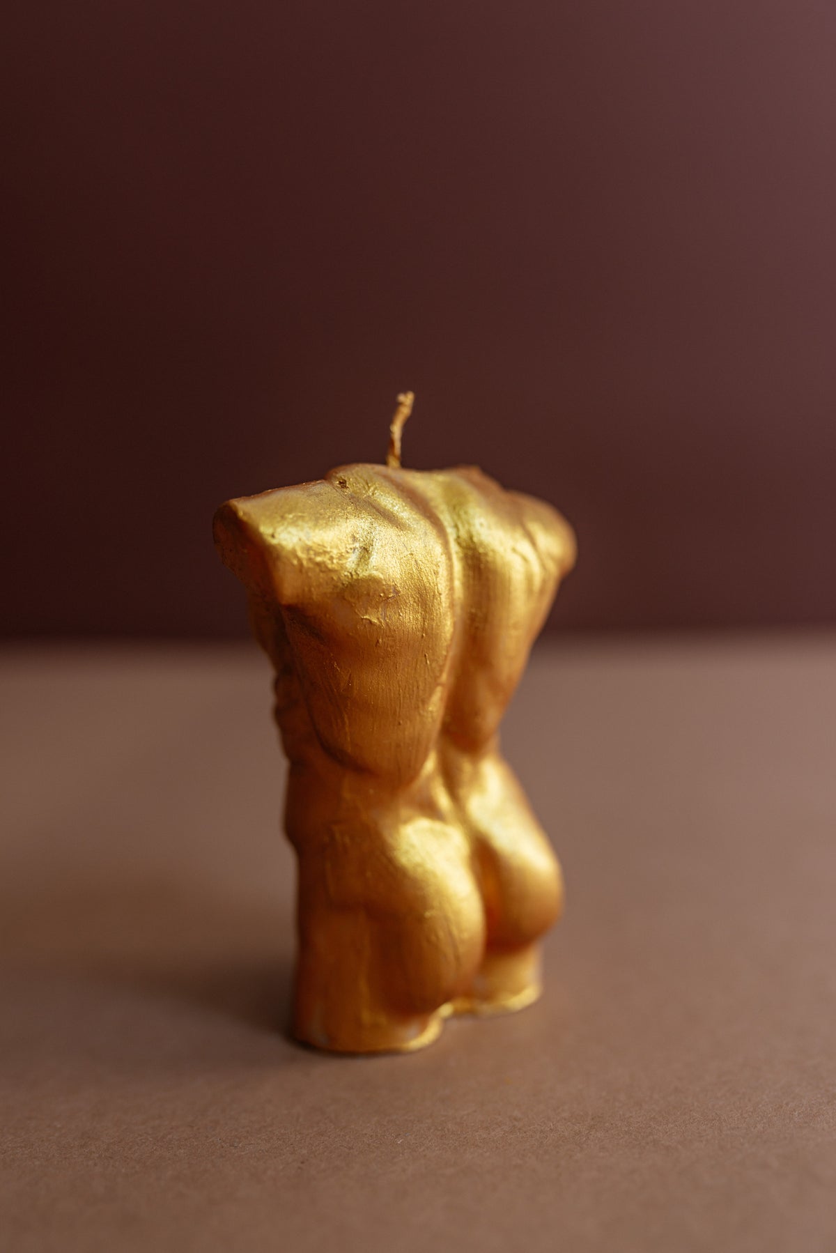 A small sculptural candle shaped like a female torso, finished in metallic gold with a single wick at the top, standing on a brown surface against a dark brown background.