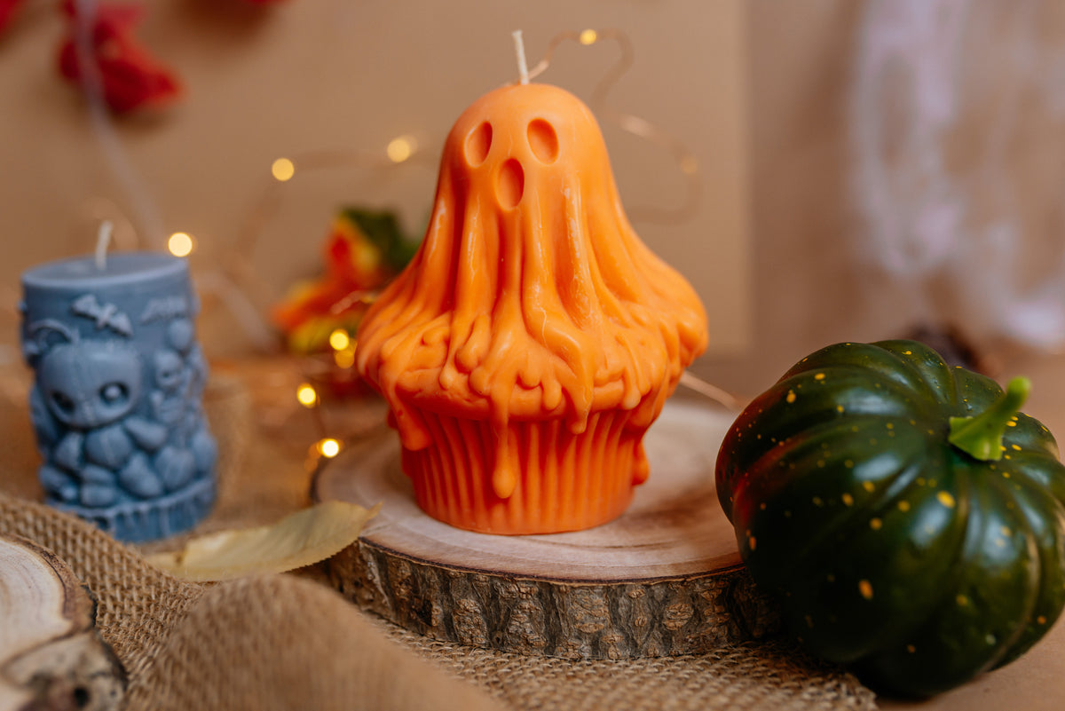 An orange ghost-shaped cupcake candle sits on a wood slice coaster, styled with a small owl candle and a green pumpkin in a warm, cozy setting.
