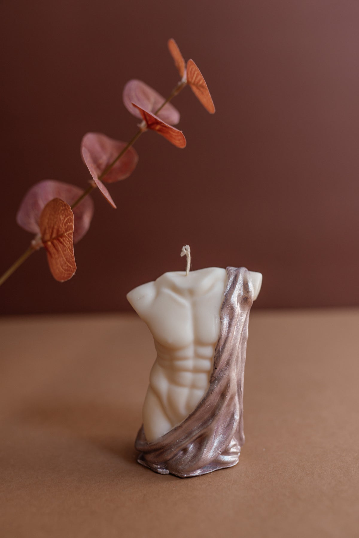 A torso-shaped candle with a cream-colored muscular chest and a draped metallic bronze fabric over one shoulder, styled on a brown surface with dried orange-brown leaves against a dark brown background.