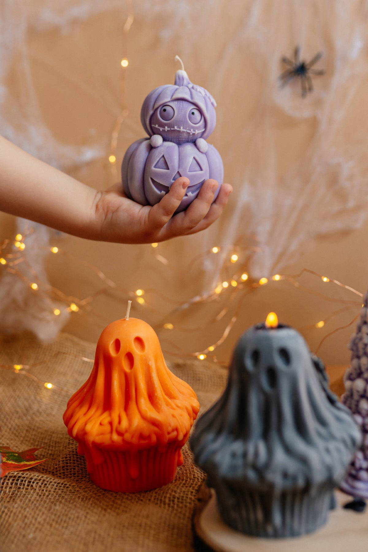 A hand holds a lavender pumpkin-shaped candle above Halloween-themed cupcakes, including an orange ghost cupcake and a gray ghost cupcake candle, with warm fairy lights and cobweb decorations in the background.