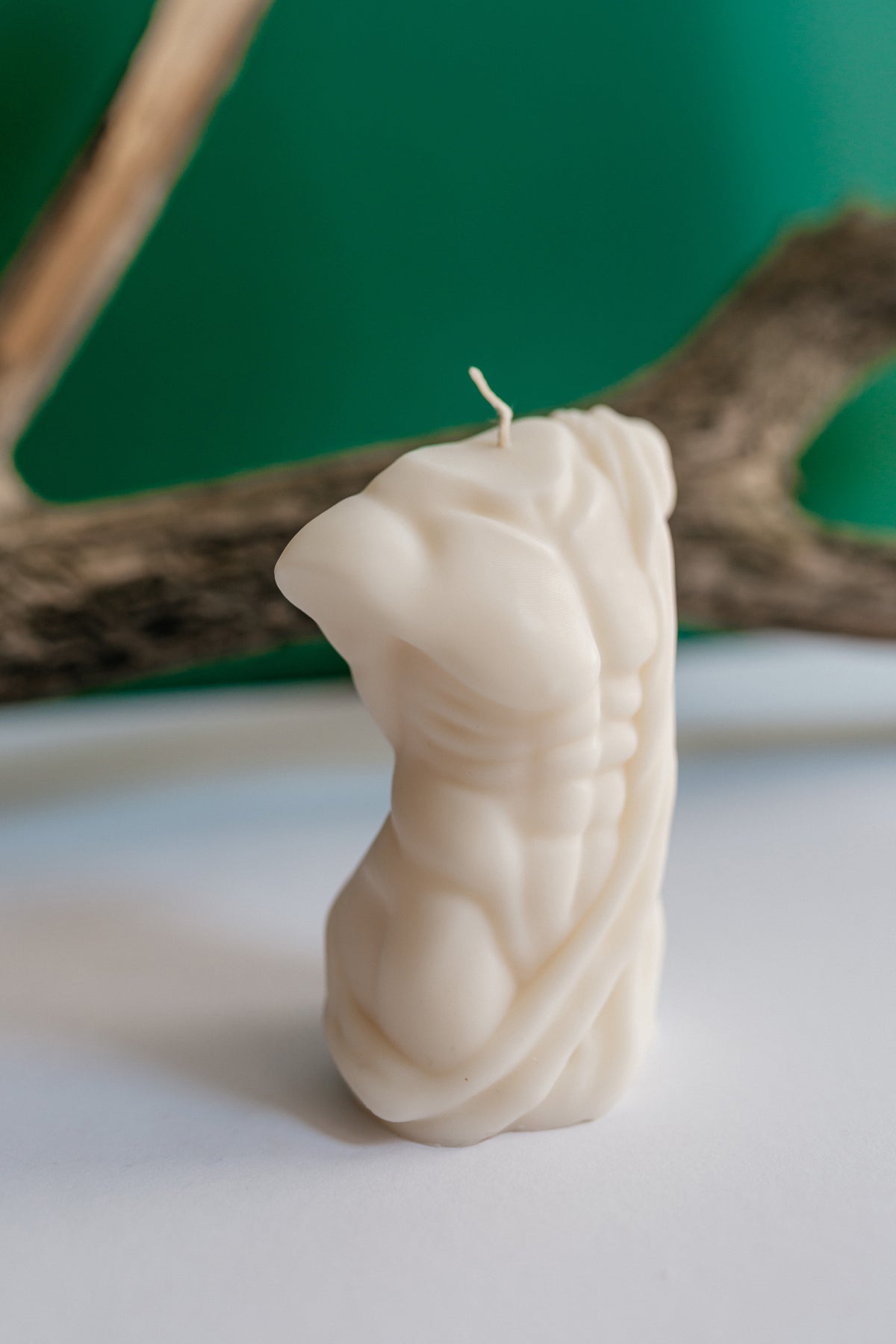 An off-white sculptural candle shaped like a muscular torso, standing upright on a white surface with a blurred green and brown background.