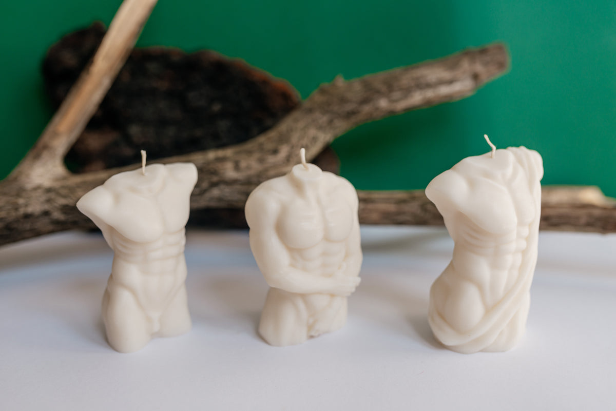 Three white sculptural candles shaped like male torso musculature, arranged in a row on a white surface with driftwood and a green backdrop.
