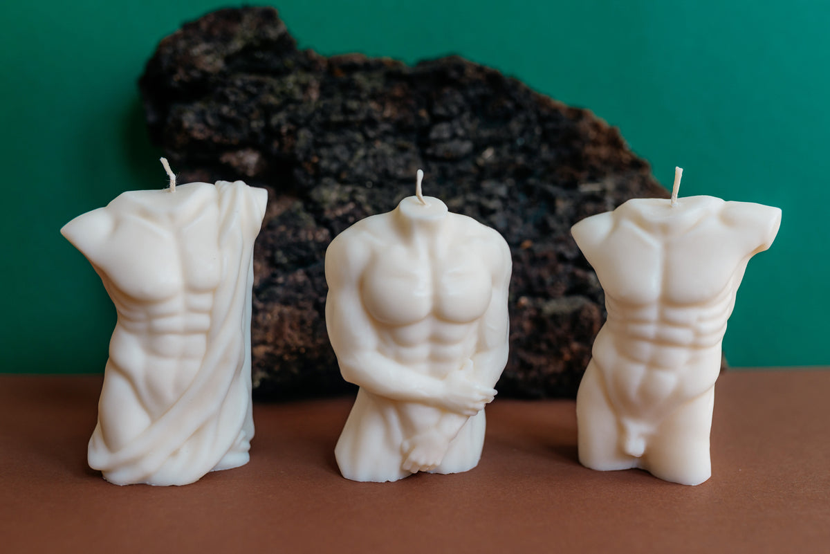 Three white torso-shaped candles displayed on a brown surface against a green background, with a dark textured rock prop behind them.