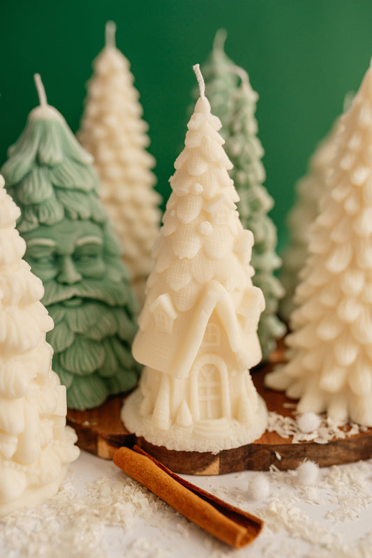 Creamy white soy wax candle shaped like a fairy tale cottage with a steep tiled roof and arched door, standing among festive winter decor.