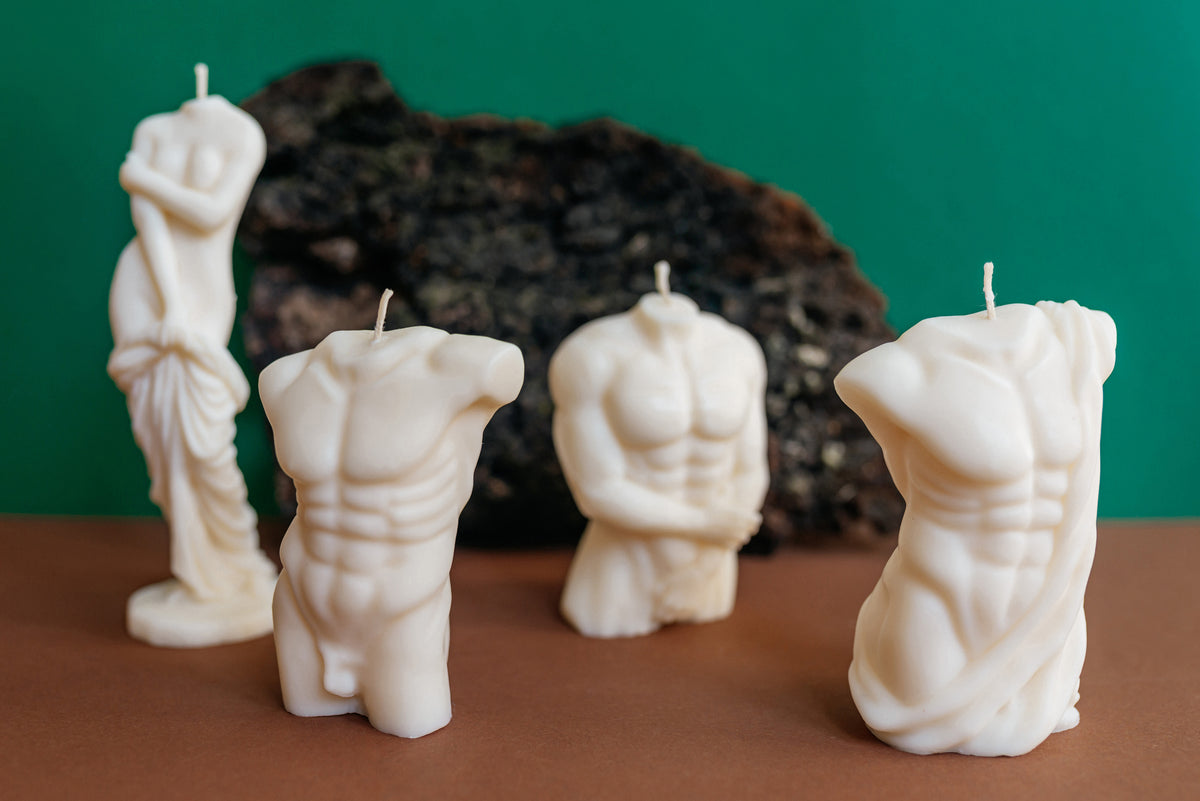 Four white sculptural candles shaped like classical male torso statues, arranged on a brown surface against a green background with a dark rock prop.