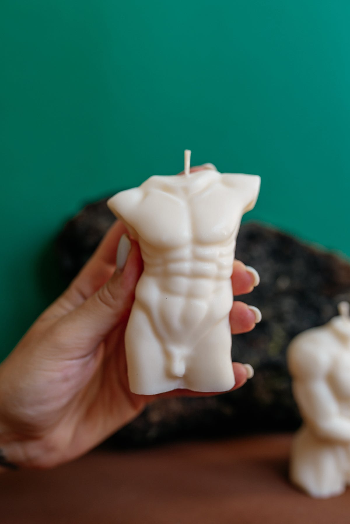 A hand holds a sculpted white body-shaped candle with a muscular torso design against a dark green background.