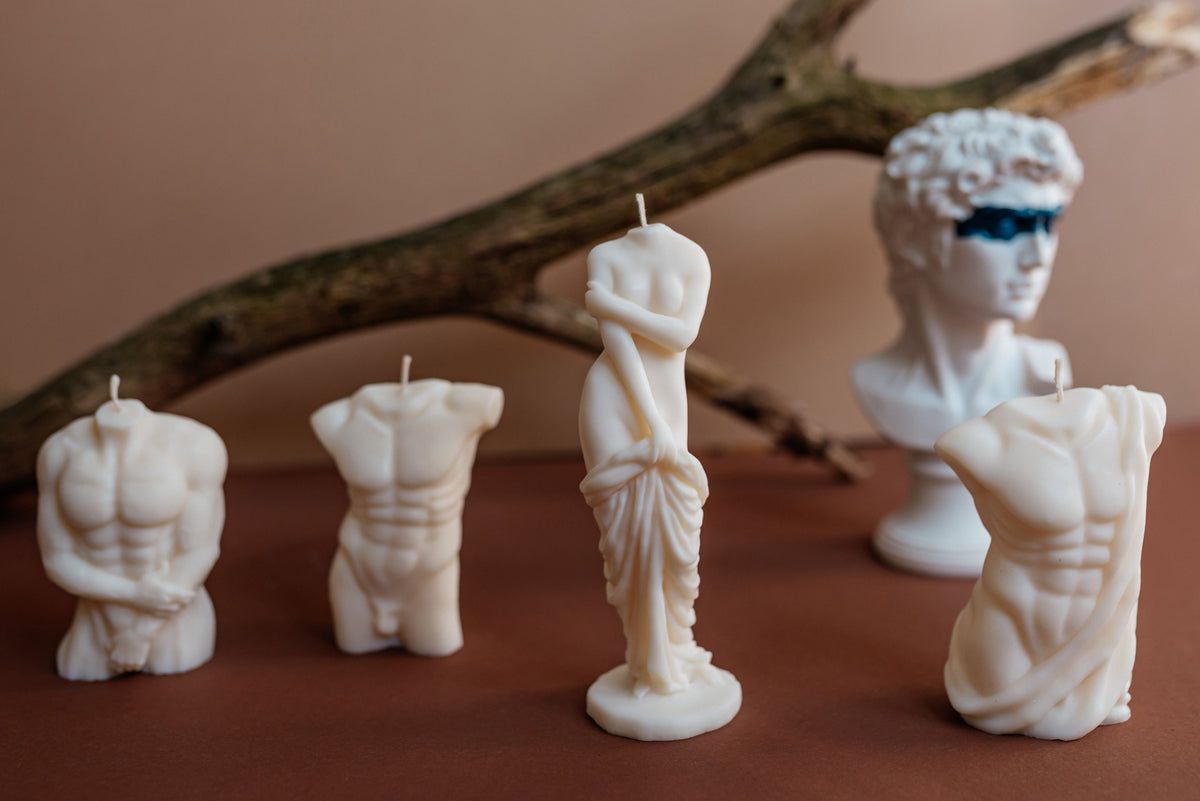 Four white sculptural candles shaped like classical male and female statues, arranged on a brown surface with a branch and a blurred bust in the background.