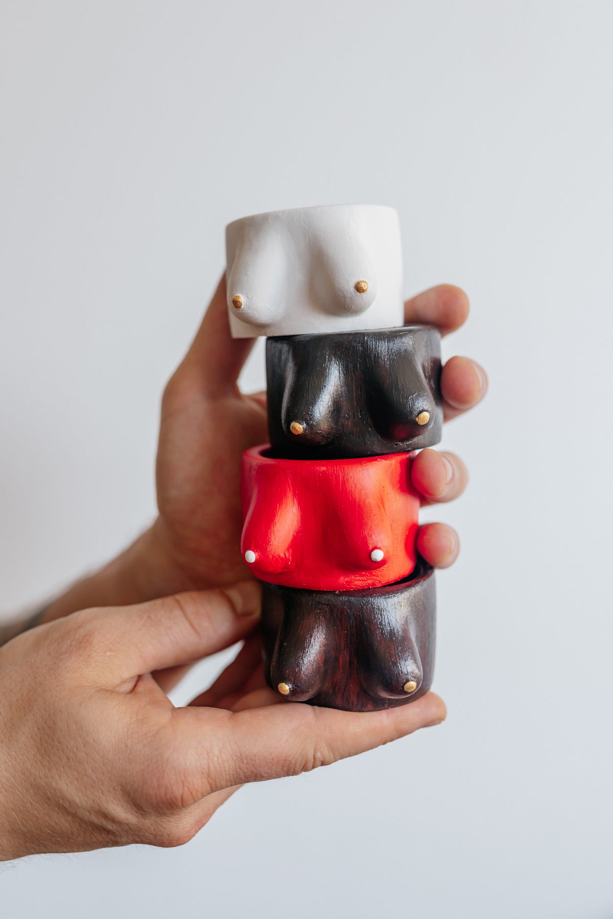 A person's hands holding a stack of three novelty ceramic cups shaped like stylized female breasts in white, black, and red, each with small gold studs, against a plain light gray background.