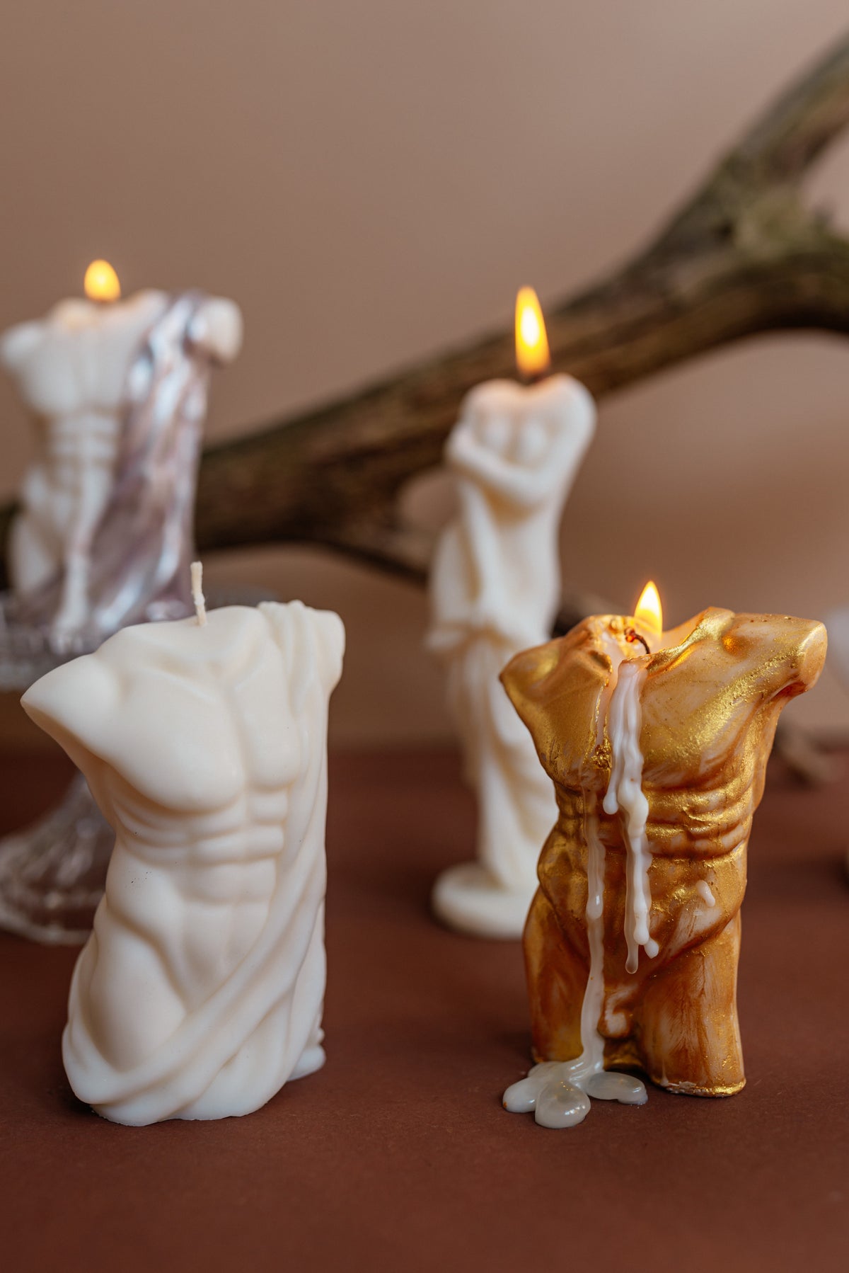 A set of sculptural candles shaped like classical male torso statues, including one white marble-style candle and one gold-toned candle with dripping wax, arranged on a brown surface with a blurred branch and warm background.