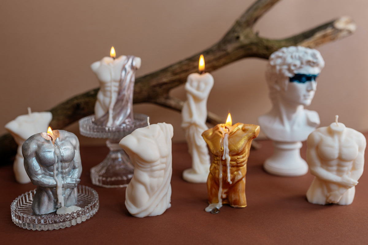 A collection of sculptural novelty candles shaped like classical Greek and Roman statues, muscular gods, and draped figures, displayed on a brown surface with a rustic branch in the background.