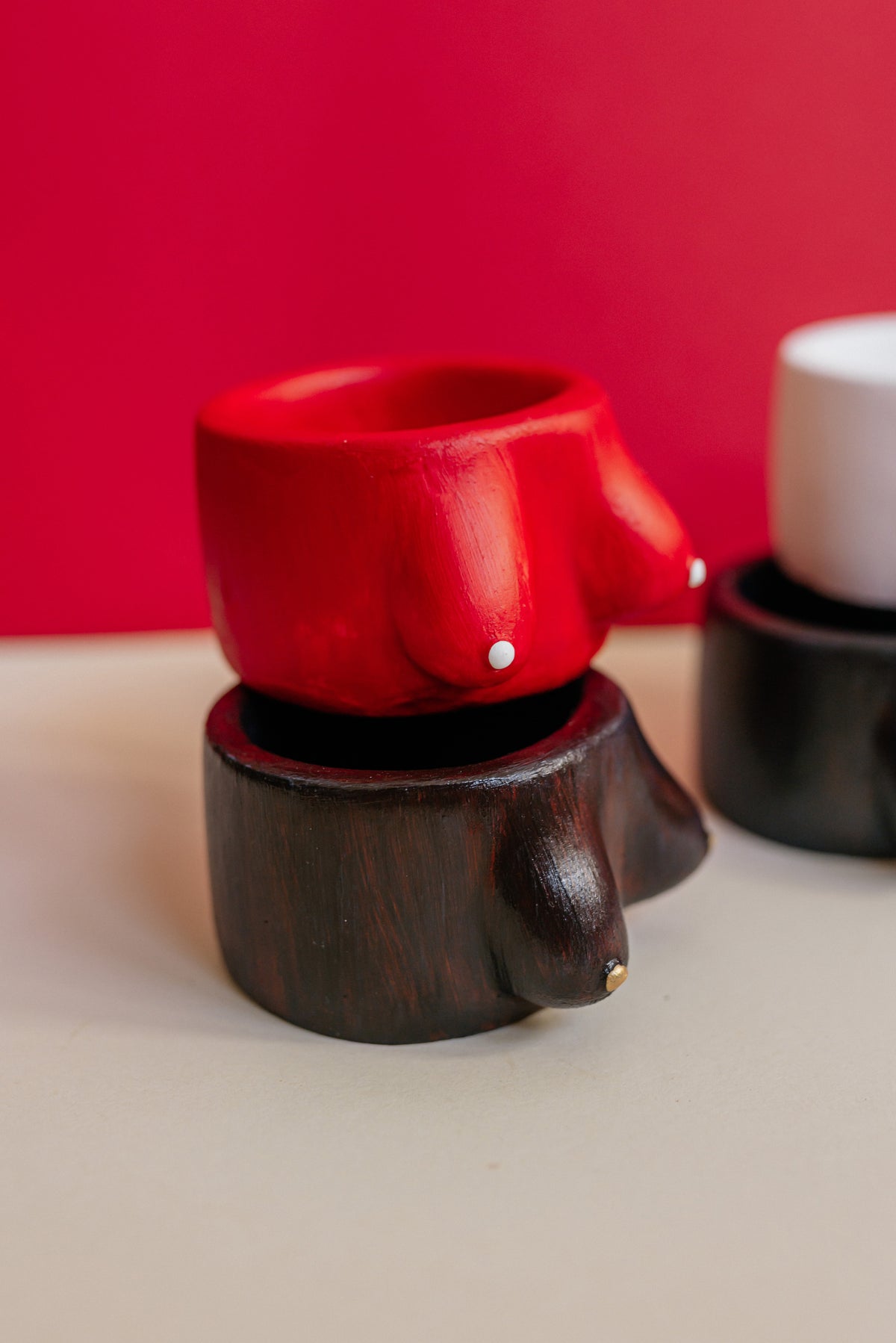A red wooden cup shaped like a male breast sits on a dark brown wooden base shaped like male genitalia, with a blurred second matching pair in the background against a red and beige backdrop.