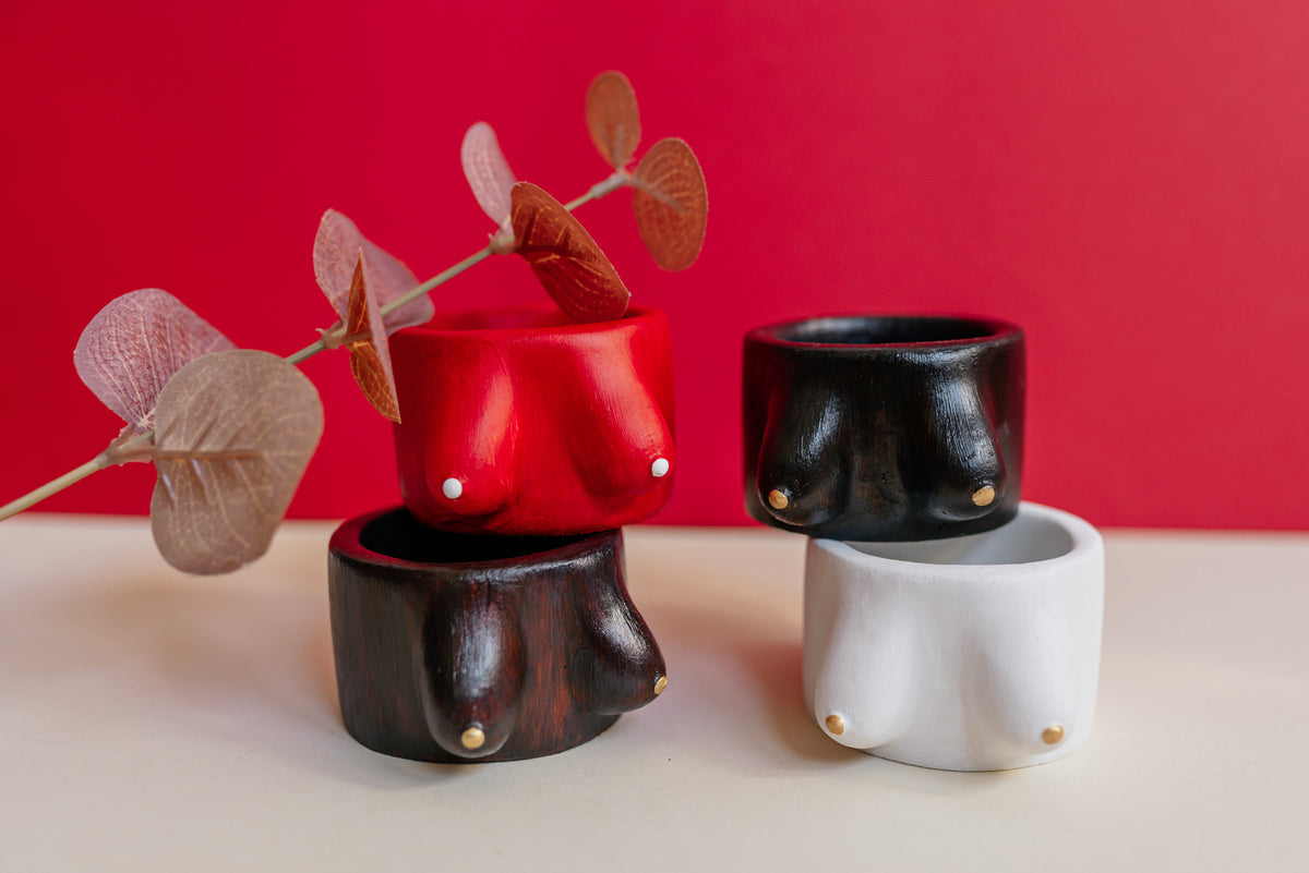Four small plaster bust-shaped containers or vases in red, black, dark brown, and white, each with a small gold stud detail, arranged on a light surface against a solid red background with dried leaves as props.