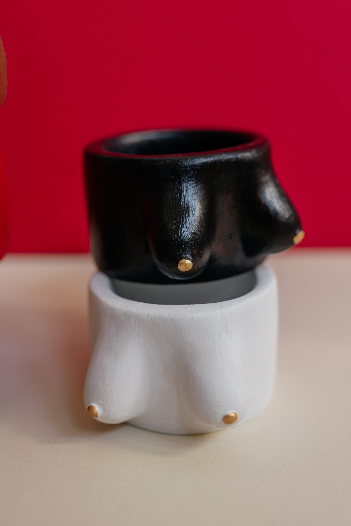 A novelty ceramic figurine shaped like a person's lower torso and upper thighs, with a glossy black top section and a matte white bottom section, accented with small gold nipple details, set against a solid red background and a beige surface.