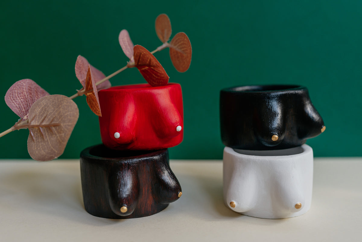 Three small decorative bust-shaped vessels in glossy red, black, and white, each with a small gold stud detail, arranged on a cream surface against a dark green background with dried pink-brown leaves in the foreground.