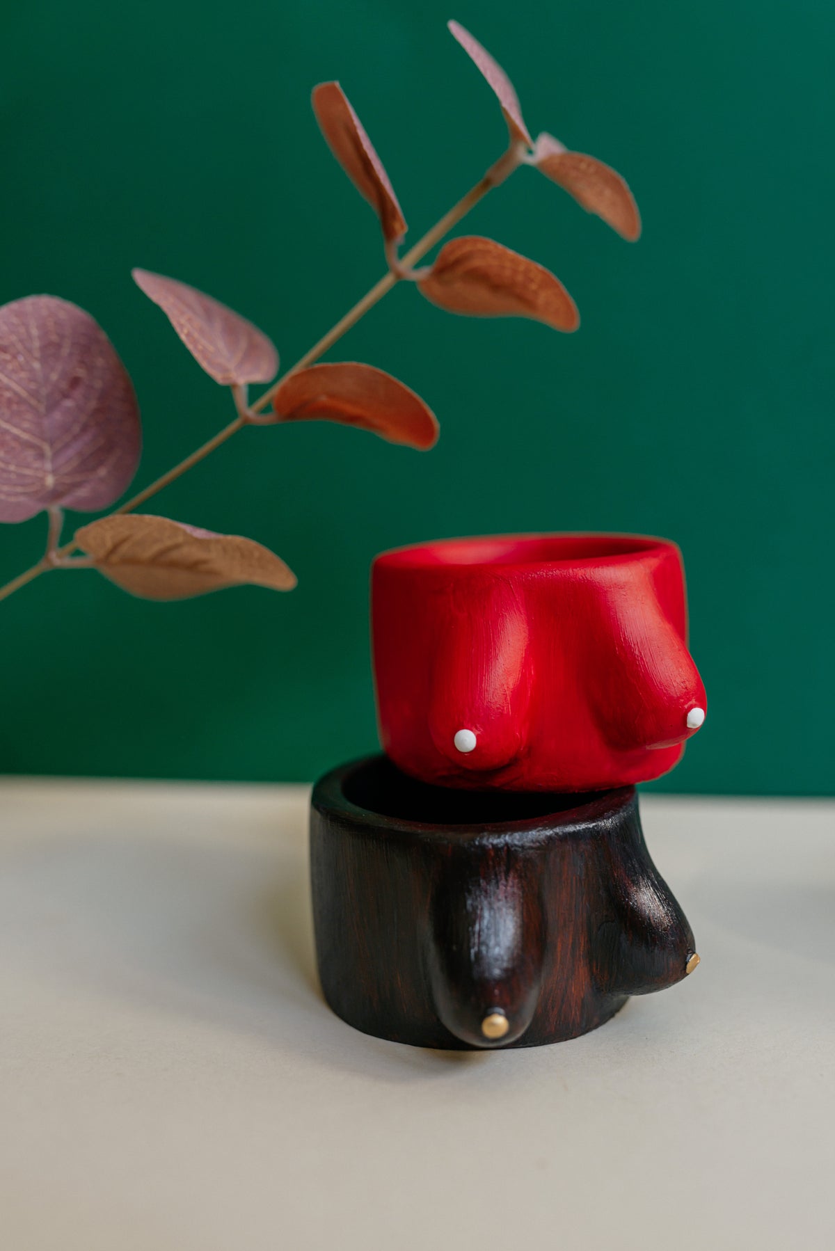 A red plaster female bust sculpture displayed on a dark wooden pedestal against a solid green background, with dried leaves in the upper left.