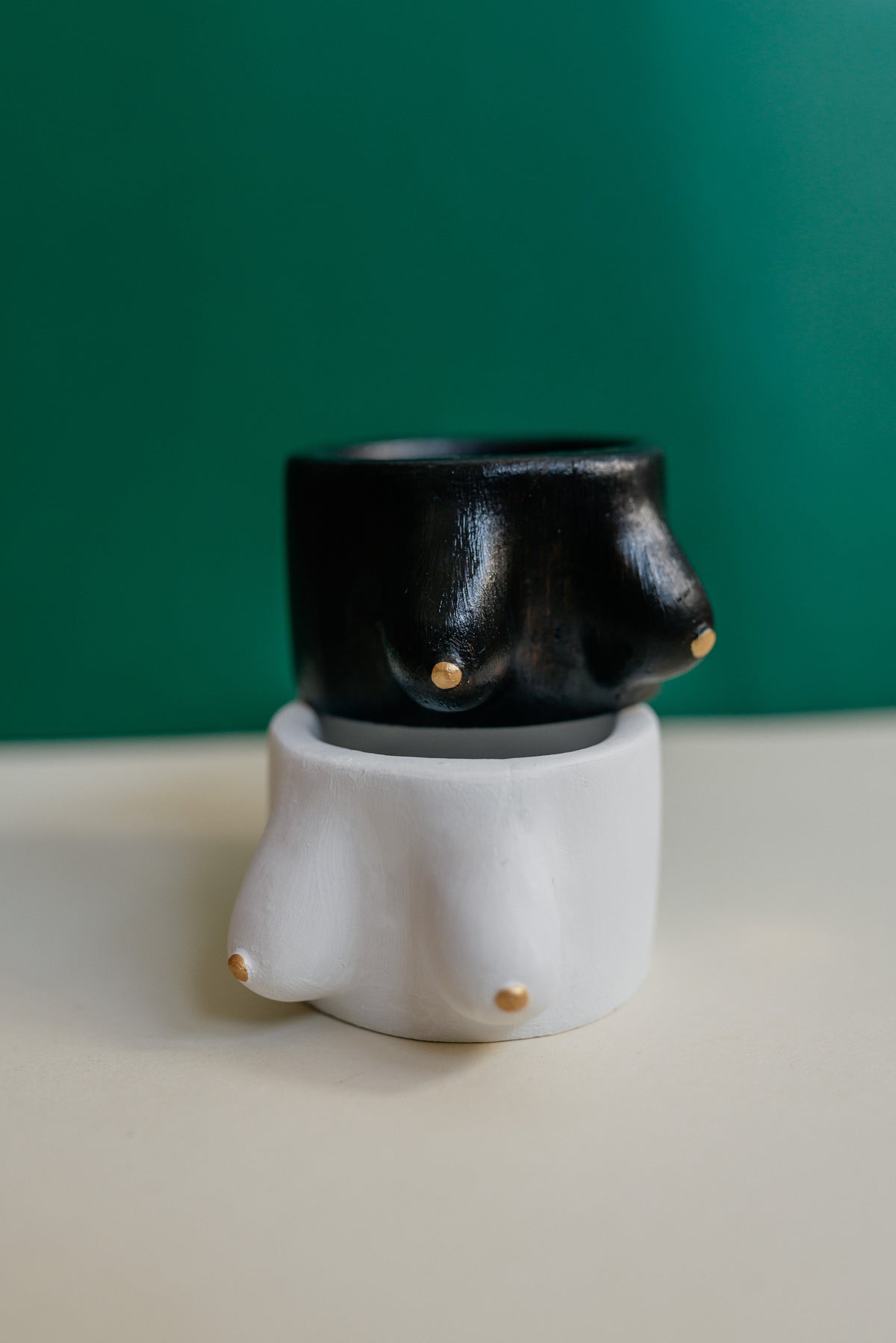 A sculptural novelty cup shaped like a stylized female breast, shown in a two-tone black and white stacked design with small gold nipple details, set against a dark green background.