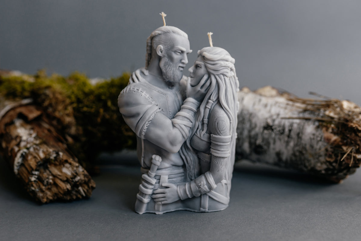 A monochrome gray sculptural candle depicting a bearded man and a woman embracing, with two small wicks on top, photographed against a soft gray background with blurred natural props.
