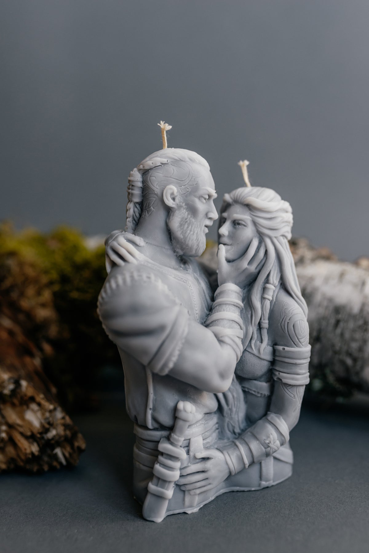 A monochrome gray sculptural candle depicting a bearded man and a woman embracing, with two small wicks on top, photographed against a soft gray background with blurred natural props.