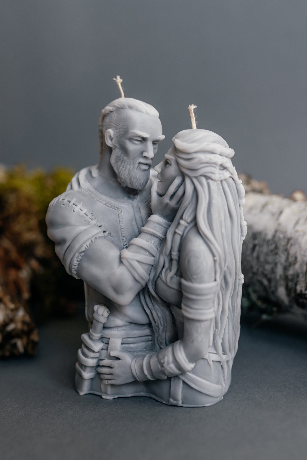 A monochrome gray sculptural candle depicting a bearded man embracing a long-haired woman, with two visible wicks, photographed against a dark gray background with blurred natural props at the sides.