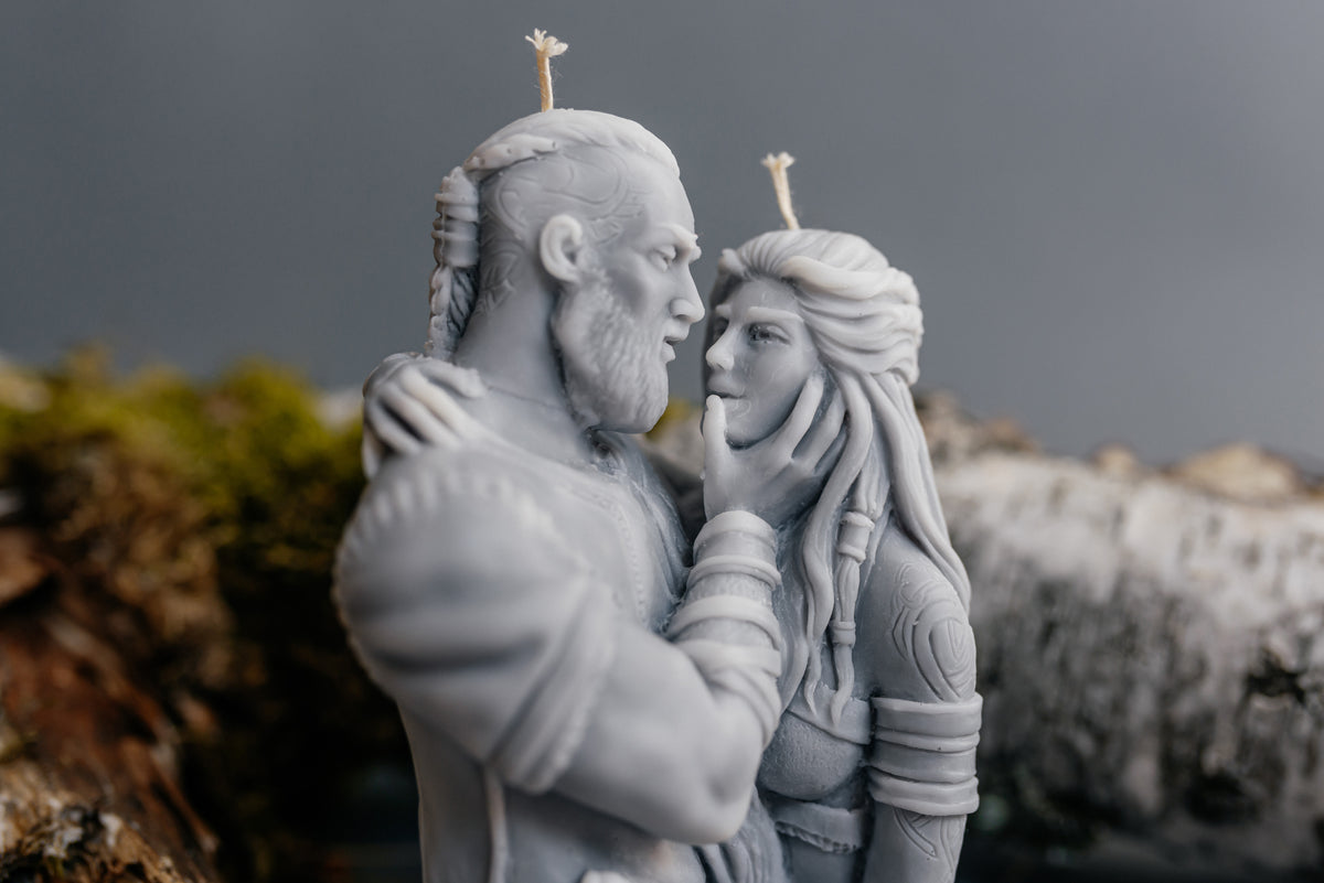 A monochrome gray sculptural candle depicting a bearded man and a woman in an embrace, with two small flame-shaped wicks visible on top, set against a blurred natural background.