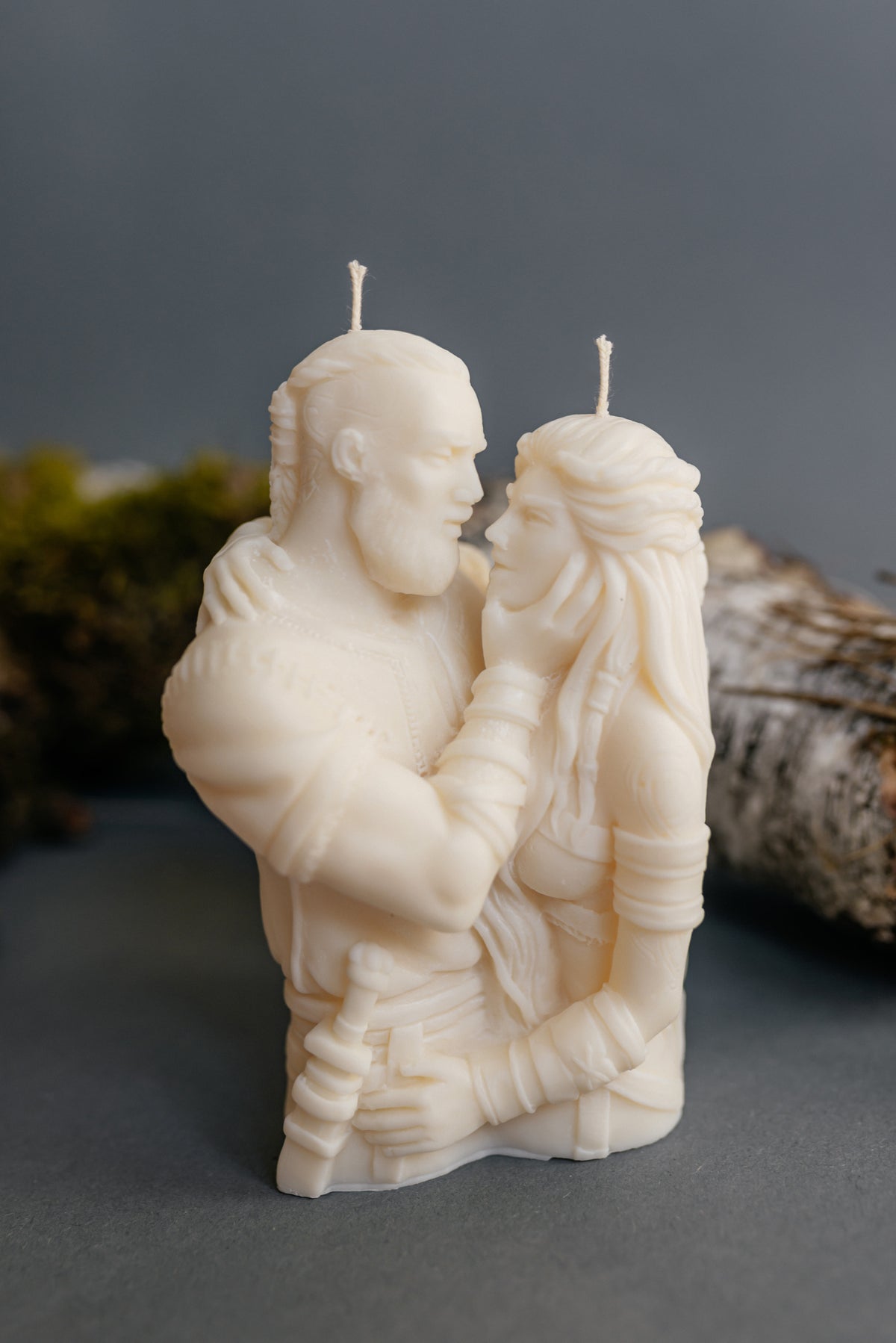 An off-white sculpted candle shaped like a bearded man and a woman embracing, photographed on a dark gray surface with blurred natural props in the background.